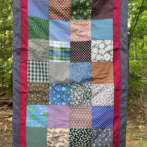 Könnte beinhalten: Ein Patchwork-Quilt mit verschiedenen gemusterten Quadraten in Blau-, Grün-, Braun- und Lilatönen. Der Quilt hat einen roten Rand und einen dunkelgrauen äußeren Rand. Der Quilt hängt draußen an einer Wäscheleine.