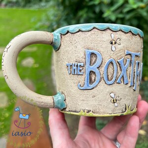 May include: A handmade ceramic mug with the text "THE BOX TREE" in blue, featuring bee illustrations. The mug has a beige base with a scalloped turquoise rim and handle.
