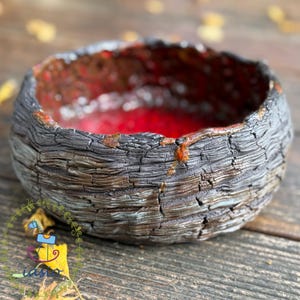 Pode incluir: Uma pequena tigela de cerâmica redonda com uma textura exterior escura e carbonizada que lembra casca de árvore. O interior da tigela apresenta um esmalte vermelho vibrante e brilhante que contrasta com a superfície exterior rugosa castanha e preta.