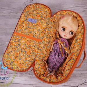以下が含まれることがあります： 花柄の裏地と人形が入った、開いたオレンジ色の人形用キャリーケース。人形はブロンドの髪で、紫色のオーバーオールを着ています。キャリーケースにはハンドルと小さな青いタグが付いています。