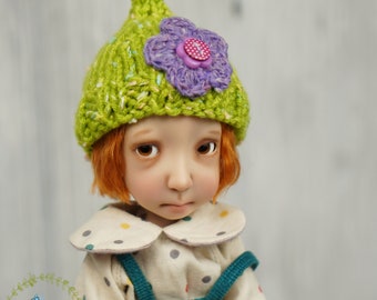 May include: A doll wearing a green and white knitted hat with a purple flower and a button on top. The doll is wearing a white shirt with colorful polka dots and blue overalls.