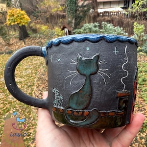 Könnte beinhalten: Handgefertigte Keramiktasse mit schwarzer Außenseite und tealfarbenem Katzendesign. Die Tasse hat einen dunkelblauen Rand und einen Henkel. Die Katze sitzt auf einer Backsteinmauer, mit einem Schornstein und Sternen.