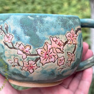 Può includere: Una tazza in ceramica con smalto screziato blu e verde acqua, caratterizzata da un disegno dipinto a mano di fiori di ciliegio rosa su rami marrone scuro. La tazza presenta un corpo arrotondato, un manico robusto e una finitura liscia e lucida.