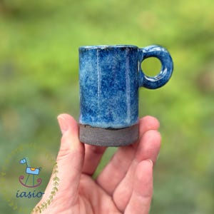 Może przedstawiać: Mały ceramiczny kubek z nakrapianą niebieską glazurą w górnej części i ciemnoszarą, teksturowaną podstawą. Kubek ma cylindryczny korpus i okrągły uchwyt wystający z boku, co podkreśla wyraźny kontrast pomiędzy dwoma rodzajami wykończenia.