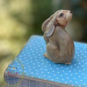 Może przedstawiać: Mała, jasnobrązowa ceramiczna figurka zająca o teksturowanej powierzchni, długich uszach i gładkim wykończeniu. Zając jest przedstawiony w pozycji siedzącej, patrząc w górę i w prawo, ukazując szczegółowe rysy twarzy i naturalną formę.