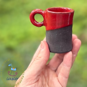 Peut inclure: Une petite tasse en céramique présentant une partie supérieure émaillée rouge vif et brillante, et une partie inférieure noire mate et texturée. La tasse possède une anse arrondie sur le côté, soulignant un contraste entre les finitions.