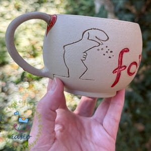 Puede incluir: Una taza de cerámica beige con superficie texturizada que presenta un dibujo de líneas grabadas en marrón oscuro de una figura y el texto pintado en rojo "foi". La taza tiene un asa curva con un pequeño acento rojo en la parte superior.