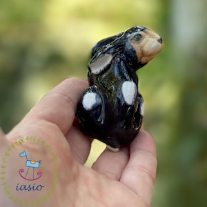 Może przedstawiać: Mała, błyszcząca ceramiczna figurka królika z czarnym tułowiem, białymi łatami na udach i jasnobrązowym pyszczkiem. Królik przedstawiony jest w pozycji siedzącej z uszami położonymi do tyłu, posiada gładkie, odblaskowe wykończenie powierzchni.