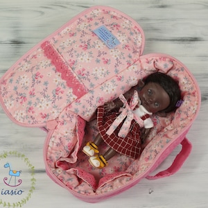 May include: A pink fabric carrying case with a floral pattern, featuring a zipper closure and lace trim, containing a small doll with dark skin and dark hair wearing a white shirt and a red plaid dress with yellow and white shoes.