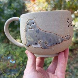 Könnte beinhalten: Ein beigefarbener Keramikbecher mit einer erhabenen, glänzend grauen Robbenillustration mit dunklen Punkten und Schnurrhaaren. Der Becher hat einen geschwungenen Henkel und eine Teilinschrift an der Seite. Die Oberfläche ist matt und gesprenkelt, und die Robbe ist im Seitenprofil mit sichtbaren Flossen dargestellt.