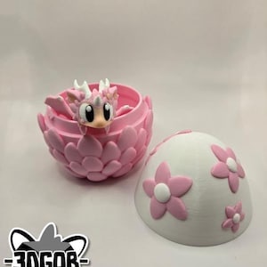 May include: A pink dragon figurine inside a pink egg-shaped container with a white egg-shaped lid with pink flowers. The lid is open and the dragon is visible inside. The container is made of plastic and has a textured surface.