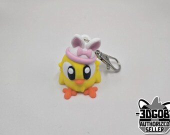 May include: A yellow chick keychain with a pink and white bunny ear hat. The keychain has a silver metal ring.