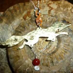 Puede incluir: Un collar de cadena de plata con un colgante de zorro de latón y una piedra preciosa roja. El zorro está saltando y tiene una textura martillada. El collar tiene una cuenta de espiral de cobre en la parte superior.