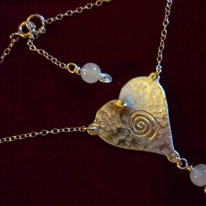 Puede incluir: Un collar de plata con un colgante en forma de coraz&oacute;n con un dise&ntilde;o en espiral y dos cuentas blancas. El collar tiene una cadena delicada.