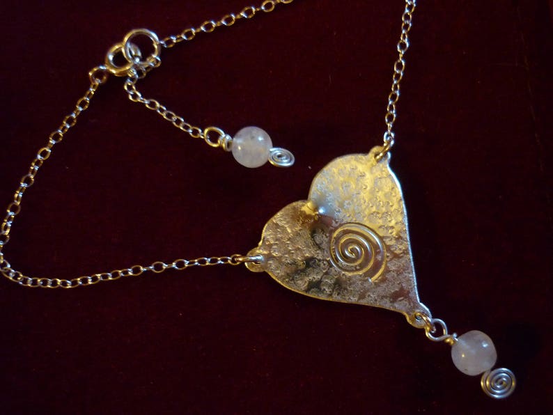Puede incluir: Un collar de plata con un colgante en forma de coraz&oacute;n con un dise&ntilde;o en espiral y dos cuentas blancas. El collar tiene una cadena delicada.