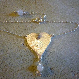 Puede incluir: Un collar de plata con un colgante en forma de coraz&oacute;n, con un dise&ntilde;o en espiral y una piedra blanca que cuelga de la parte inferior.