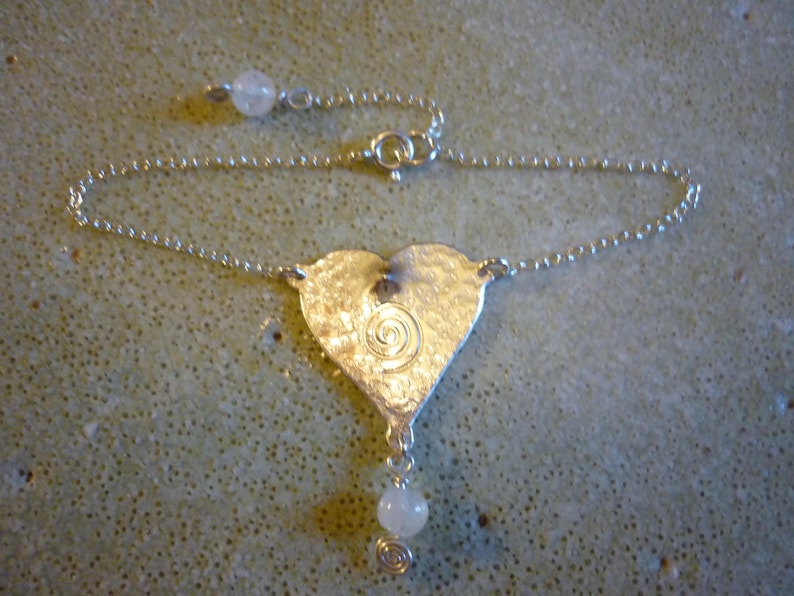 Puede incluir: Un collar de plata con un colgante en forma de coraz&oacute;n, con un dise&ntilde;o en espiral y una piedra blanca que cuelga de la parte inferior.