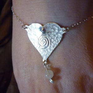 Puede incluir: Un brazalete de plata con un dije en forma de coraz&oacute;n con un dise&ntilde;o en espiral y una cuenta blanca.
