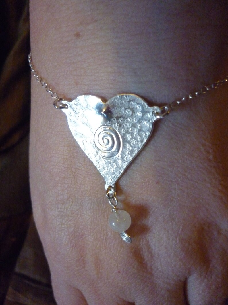 Puede incluir: Un brazalete de plata con un dije en forma de coraz&oacute;n con un dise&ntilde;o en espiral y una cuenta blanca.