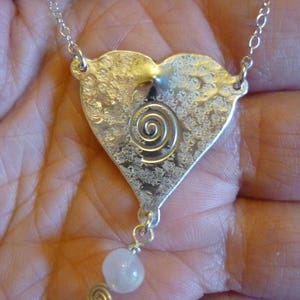 Puede incluir: Un collar con un colgante en forma de coraz&oacute;n de plata con un dise&ntilde;o en espiral y una sola perla blanca colgando de la parte inferior.