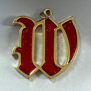 Puede incluir: Un colgante dorado con forma de la letra "W". El interior de la letra está relleno de esmalte rojo vibrante. El colgante tiene un lazo en la parte superior para sujetarlo a una cadena o collar.