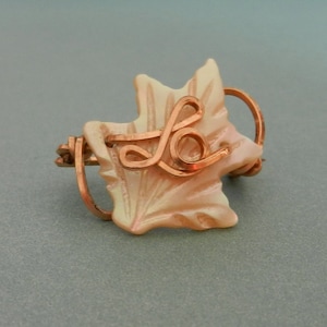 Könnte beinhalten: Ein Ring aus Kupferdraht mit einem zarten, weißen, blattartigen Anhänger. Der Anhänger hat ein Kupferdrahtdesign in Form des Buchstabens "L" darauf.