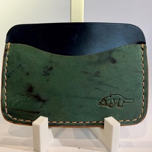 May include: A handcrafted leather cardholder in green and black. The green section has a distressed look with a small embossed platypus design. The edges are stitched with a light brown thread. The cardholder is displayed on a white stand.