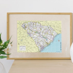 K&ouml;nnte beinhalten: Gerahmte Karte von South Carolina mit detaillierten Stra&szlig;ennetzen, St&auml;dten und geografischen Merkmalen. Die Karte befindet sich in einem hellen Rahmen, mit einer wei&szlig;en Vase und Blumen im Vordergrund. Vintage-Stil.