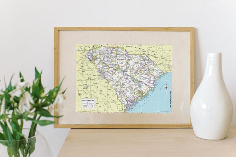 K&ouml;nnte beinhalten: Gerahmte Karte von South Carolina mit detaillierten Stra&szlig;ennetzen, St&auml;dten und geografischen Merkmalen. Die Karte befindet sich in einem hellen Rahmen, mit einer wei&szlig;en Vase und Blumen im Vordergrund. Vintage-Stil.