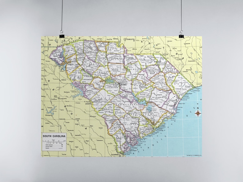 K&ouml;nnte beinhalten: Eine detaillierte Karte von South Carolina mit den County-Grenzen, St&auml;dten und geografischen Merkmalen. Die Karte ist auf einem hellgelben Hintergrund mit einem blauen Ozeanbereich gedruckt. Die Karte wird von schwarzen Clips gehalten.