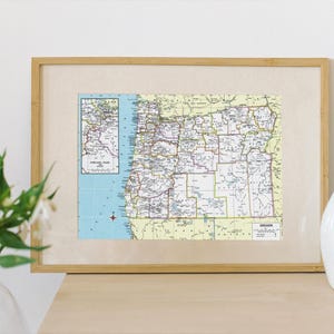 May include: A framed vintage map of Oregon with a light brown wooden frame. The map is in shades of green, yellow, and blue. The map shows the state's counties and major cities. The text "OREGON" is printed in the bottom right corner of the map.