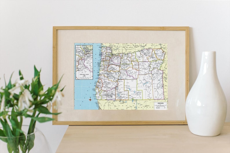 May include: A framed vintage map of Oregon with a light brown wooden frame. The map is in shades of green, yellow, and blue. The map shows the state's counties and major cities. The text "OREGON" is printed in the bottom right corner of the map.