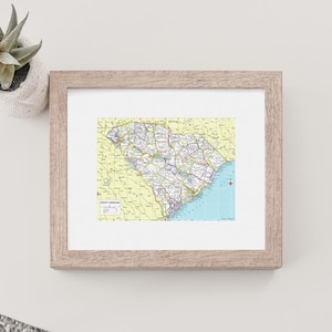 K&ouml;nnte beinhalten: Gerahmte Karte von South Carolina mit detaillierter Darstellung der Stra&szlig;en, St&auml;dte und geografischen Merkmale des Bundesstaates. Die Karte hat einen gelben Hintergrund mit blauen K&uuml;stenbereichen. Der Rahmen ist hellbraun. Eine kleine Sukkulente in einem wei&szlig;en Topf ist sichtbar.