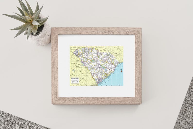 K&ouml;nnte beinhalten: Gerahmte Karte von South Carolina mit detaillierter Darstellung der Stra&szlig;en, St&auml;dte und geografischen Merkmale des Bundesstaates. Die Karte hat einen gelben Hintergrund mit blauen K&uuml;stenbereichen. Der Rahmen ist hellbraun. Eine kleine Sukkulente in einem wei&szlig;en Topf ist sichtbar.