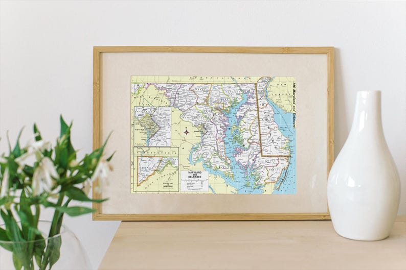 May include: A framed vintage map of Maryland and Delaware with a light yellow background and blue and green accents. The map shows major roads and cities.