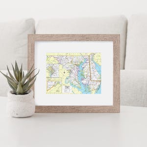 May include: A framed map of Maryland with a light wood frame. The map is in a vintage style with blue and green colors. The map shows the state's major cities and highways.