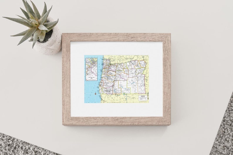 May include: A framed map of Oregon with a smaller inset map of the state's coastline. The map is printed in shades of yellow, green, and blue, with black lines indicating roads and borders. The frame is a light wood colour.