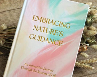 May include: A hardcover journal titled "Embracing Nature's Guidance" with a pastel pink, teal, and cream cover. The book's title is in gold lettering. The subtitle reads "An Interactive Journey Through the Seasons of Life."