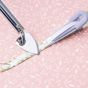 May include: A close-up of a silver pressing tool with a triangular head and a purple fabric pressing tool. Both tools are being used to press a white and yellow fabric strip.