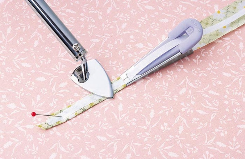 May include: A close-up of a silver pressing tool with a triangular head and a purple fabric pressing tool. Both tools are being used to press a white and yellow fabric strip.