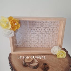 May include: A rectangular wooden box with a clear front panel and a geometric patterned background. The box is decorated with yellow and white fabric flowers. The text "L'atelier d'Evelyna" is visible on the wooden surface.