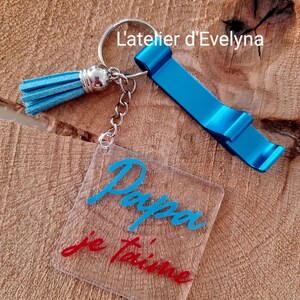Pode incluir: Um porta-chaves abridor de garrafas azul com uma borla e um pingente quadrado transparente. O pingente tem as palavras "Papa je t'aime" em escrita azul e vermelha. O porta-chaves tamb&eacute;m tem o texto "L'atelier d'Evelyna".