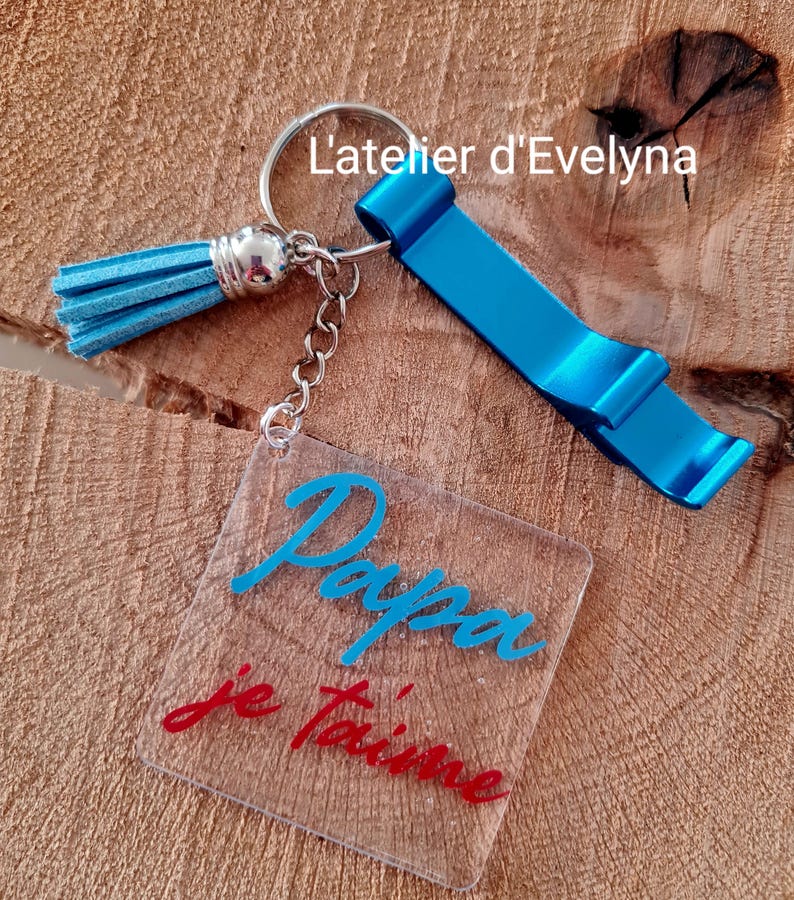 Pode incluir: Um porta-chaves abridor de garrafas azul com uma borla e um pingente quadrado transparente. O pingente tem as palavras "Papa je t'aime" em escrita azul e vermelha. O porta-chaves tamb&eacute;m tem o texto "L'atelier d'Evelyna".
