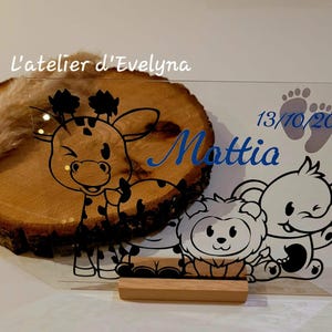 Peut inclure: Une tranche de bois avec une illustration de girafe et un panneau acrylique transparent avec un lion, un éléphant et le nom "Mattia" en bleu. Le panneau comprend également la date 13/10/2025 et des empreintes de pas. Le texte "l'atelier d'Evelyna" est également présent.