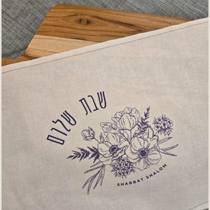 Könnte beinhalten: Ein Leinen-Tischläufer mit floralem Design und den Worten "Shabbat Shalom" in Lila. Das florale Design umfasst detaillierte Strichzeichnungen von Blumen und Blättern. Der Läufer liegt auf einem Holzschneidebrett.