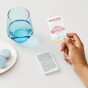 K&ouml;nnte beinhalten: Ein klares, stieloses Glas mit blauer Fl&uuml;ssigkeit steht neben einem wei&szlig;en Teller mit zwei hellblauen Makronen. Eine Karte mit dem Text "Traveler's Prayer" und einer Strandillustration wird von einer Hand gehalten.