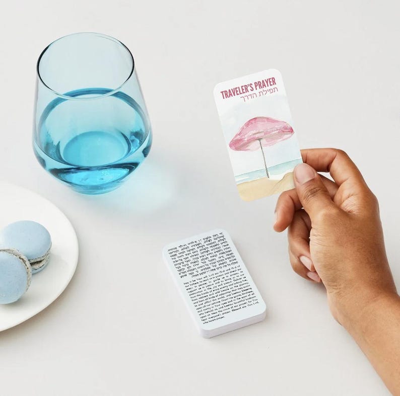 K&ouml;nnte beinhalten: Ein klares, stieloses Glas mit blauer Fl&uuml;ssigkeit steht neben einem wei&szlig;en Teller mit zwei hellblauen Makronen. Eine Karte mit dem Text "Traveler's Prayer" und einer Strandillustration wird von einer Hand gehalten.