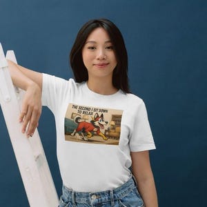 May include: White t-shirt featuring a cartoon dog in a red jacket, with the text "The second I sit down to relax." The dog is running towards a couch. The model is leaning on a white ladder against a blue background.
