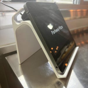 May include: A black tablet displaying "Parkway Pizza" in white text, mounted on a white and gray stand. The stand is on a stainless steel surface. A black cord is connected to the tablet.
