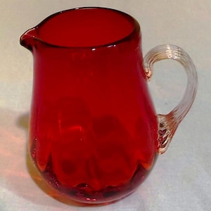 May include: A vibrant red glass pitcher with a clear, twisted handle. The pitcher has a rounded base and a pouring spout. The glass appears to be a deep, rich red color, with a slightly textured appearance near the base.